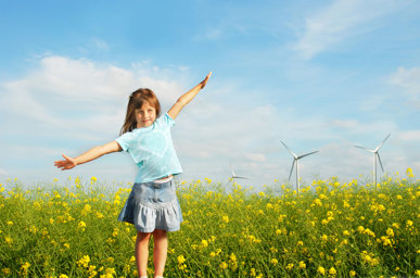 Flicka står med utsträckta armar i ett rapsfält med vindkraftverk bakom