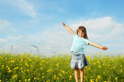 Flicka står med utsträckta armar i ett rapsfält med vindkraftverk bakom