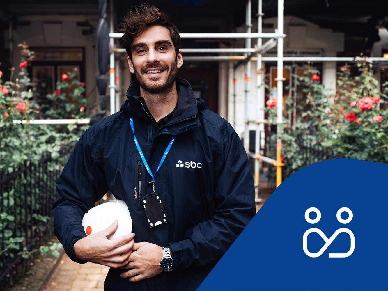 SBCs byggprojektledare Patrik står framför en fastighet med byggnadsställning och rosenrabatt. Projektledningsikon.