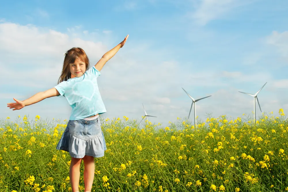 Flicka står med utsträckta armar i ett rapsfält med vindkraftverk bakom.