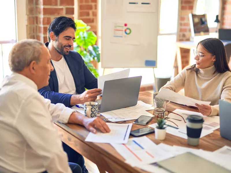 Tre personer sitter samlade runt ett mötesbord med datorer och papper