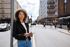 tjej står utomhus lutande mot en stolpe med mobilen i handen och kollar ut över storstaden