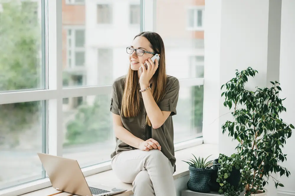 Ung kvinna sitter med sin laptop vid fönstret i sin bostadsrättslägenhet och talar i mobiltelefon.
